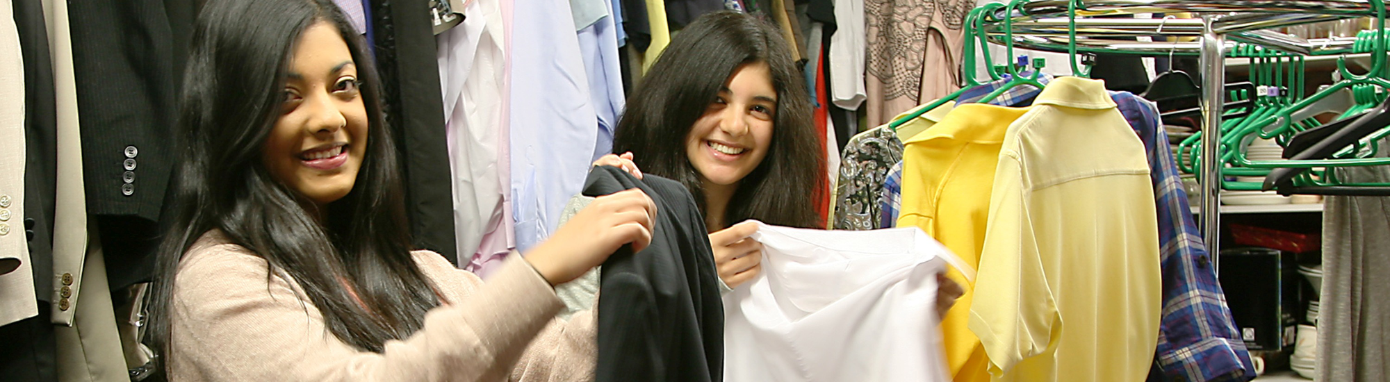 An image of two young women smiling at the camera whilst sorting clothes in a charity shop